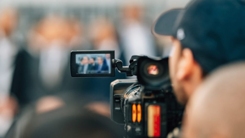 Professional video camera filming a live event, its screen showing the stage